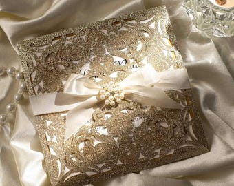May include: Gold glitter laser cut wedding invitation with a white satin ribbon and pearl embellishment. The invitation features a delicate floral design.