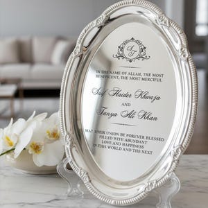 May include: An oval silver-toned decorative tray with an ornate, textured rim. The center features black printed text: In the name of Allah, the most beneficent, the most merciful, Saif Haider Khwaja and Tanya Ali Khan, may their union be forever blessed.