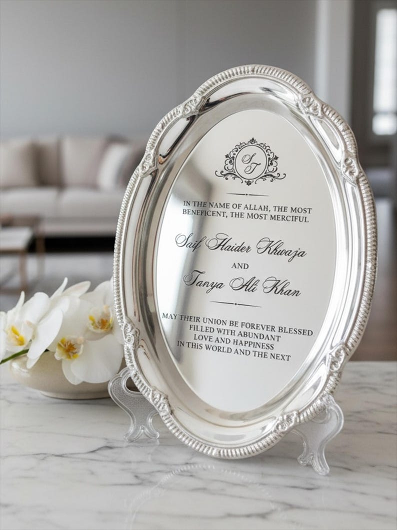 May include: An oval silver-toned decorative tray with an ornate, textured rim. The center features black printed text: In the name of Allah, the most beneficent, the most merciful, Saif Haider Khwaja and Tanya Ali Khan, may their union be forever blessed.