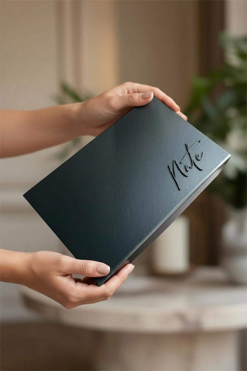 以下が含まれることがあります： 「Note」の文字がエンボス加工された、濃いティール色の長方形の箱。箱は両手で持たれています。背景はぼやけており、緑とニュートラルな色調のインテリアがかすかに見えます。