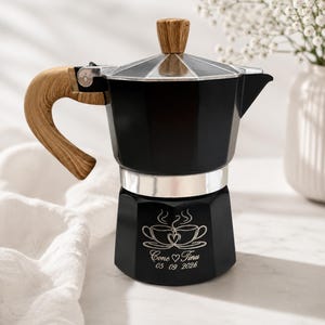 Puede incluir: Una cafetera moka negra con mango y pomo de tapa con efecto madera. La base presenta un grabado de dos tazas de café con un corazón, los nombres Cone y Tinu, y la fecha 05 09 2026. El cuerpo de metal tiene un diseño facetado.