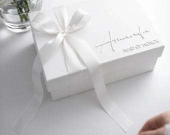 Könnte beinhalten: Eine weiße Geschenkbox mit weißem Satinband und der Aufschrift "Amanda, Maid of Honor". In der Nähe befindet sich ein Strauß weißer Rosen. Das Design ist sauber und elegant, ideal für besondere Anlässe.