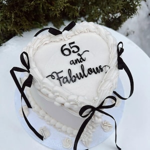 Puede incluir: Un pastel blanco en forma de corazón decorado con glaseado blanco, lazos negros y el texto "65 and Fabulous". El pastel está sobre un plato blanco, con un fondo nevado y ramas de hoja perenne.