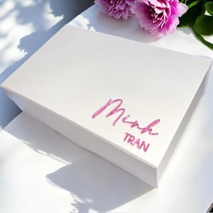 May include: A white gift box with a pink, cursive name and the last name "TRAN" in a clear, modern font. The box is rectangular and sits on a white surface, with pink flowers in the background.