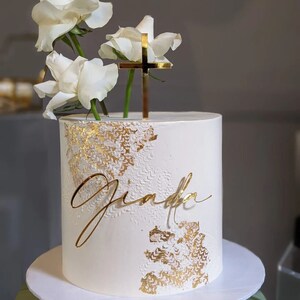 May include: White cake with gold leaf accents and a gold cross topper. The cake is decorated with white roses and the name "Giada" is written in gold script.
