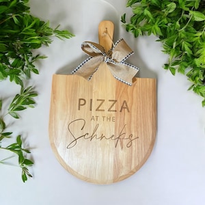 Peut inclure: Une pelle à pizza en bois clair ornée d'un nœud en toile de jute beige avec une bordure à carreaux noirs et blancs attachée au manche. La surface est gravée avec le texte PIZZA AT THE Schneks en lettres capitales et cursives.