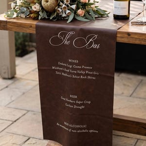 May include: A dark brown leather-like table runner featuring white printed text that reads The Bar, WINES, Umberto Luigi Cosmo Prosecco, Wickham's Road Yarra Valley Pinot Gris, Torzi Matthew's Schist Rock Shiraz, BEER, Great Northern Super Crisp, Carlton Draught, NON ALCOHOLIC, An assortment of non-alcoholic options.