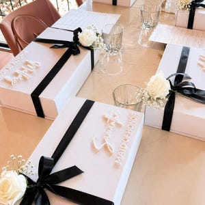 May include: White gift boxes with black ribbon and floral accents are arranged on a table. Some boxes have names like "Samantha" and "Maid of Honor" in white script. Clear glasses and a pink chair are also visible.