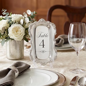 May include: A rectangular silver-toned table number sign featuring an ornate, embossed frame and a white center insert with the black text Table 4 and a decorative horizontal line element, displayed upright on a clear plastic stand.
