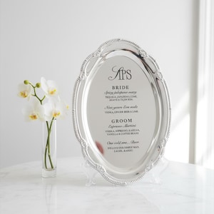 May include: A silver oval tray with a decorative border, displaying a wedding cocktail menu. The menu lists drink options for the bride and groom, along with a floral arrangement in a clear vase.