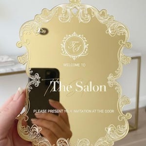 Peut inclure: Un panneau miroir doré avec un bord festonné orné et des détails gravés en blanc. Le texte indique : "WELCOME TO The Salon" et "PLEASE PRESENT YOUR INVITATION AT THE DOOR," avec un monogramme circulaire central portant les lettres "TS."