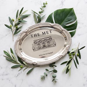 May include: An oval silver-toned metal tray with a decorative beaded rim. The center features a black line drawing of The Met museum building with the text "THE MET" above and "Where We Got Engaged" printed below the illustration.