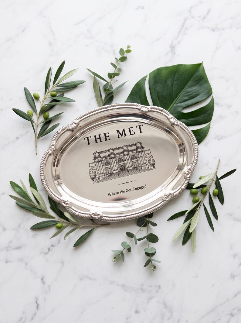 May include: An oval silver-toned metal tray with a decorative beaded rim. The center features a black line drawing of The Met museum building with the text "THE MET" above and "Where We Got Engaged" printed below the illustration.