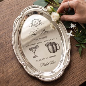 May include: An oval silver-toned serving tray with an ornate embossed border. The center features black printed text reading The Bar, Our favorites, CLASSIC MARGARITA, MOSCOW MULE, WINE, BEER & SPIRITS, Hosted Bar, and illustrations of a cocktail glass and mug.