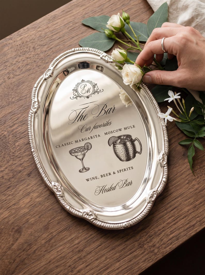 May include: An oval silver-toned serving tray with an ornate embossed border. The center features black printed text reading The Bar, Our favorites, CLASSIC MARGARITA, MOSCOW MULE, WINE, BEER & SPIRITS, Hosted Bar, and illustrations of a cocktail glass and mug.