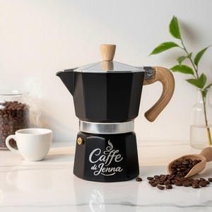 May include: Black stovetop espresso maker with a wooden handle and lid knob. The words "Caffe di Jenna" are printed on the front. Coffee beans and a white cup are in the background. A wooden scoop holds more coffee beans.