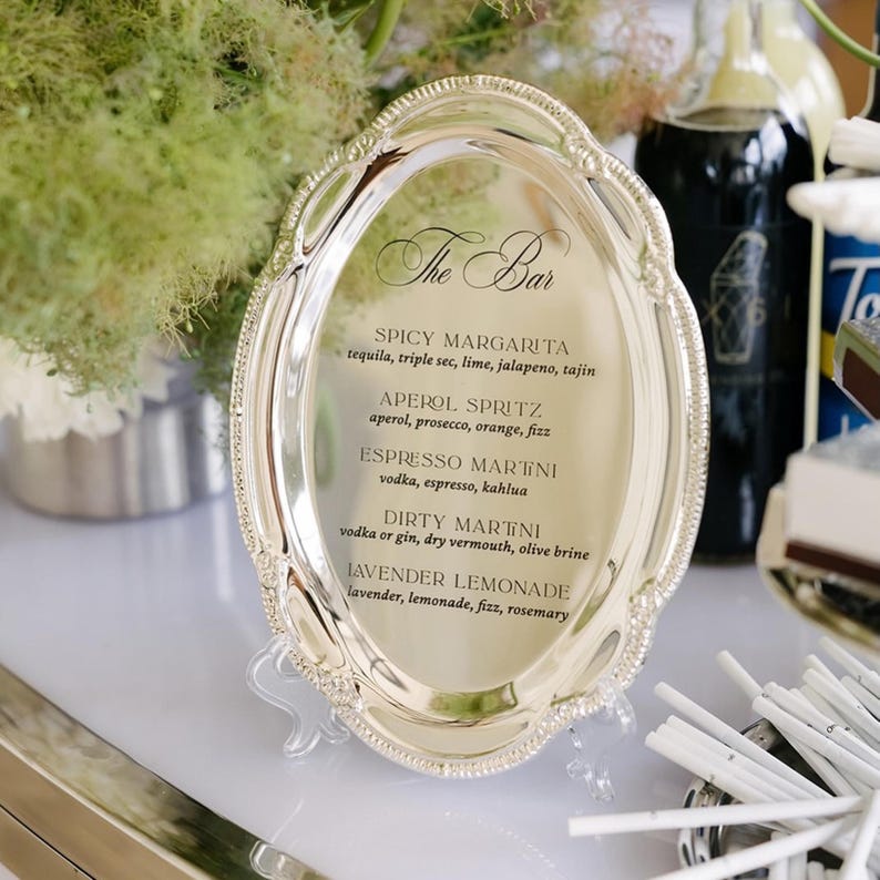 May include: An oval silver-toned serving tray used as a bar menu sign. The tray features an ornate, beaded rim and displays black printed text listing cocktail options including Spicy Margarita, Aperol Spritz, Espresso Martini, and Dirty Martini.