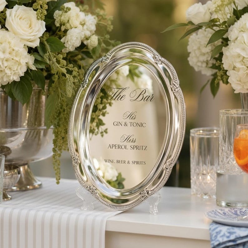 May include: An oval silver-toned mirror with an ornate beaded border, displaying black text that reads: The Bar, His GIN & TONIC, Hers APEROL SPRITZ, WINE, BEER & SPIRITS. The mirror is supported by a clear acrylic stand on a white surface.