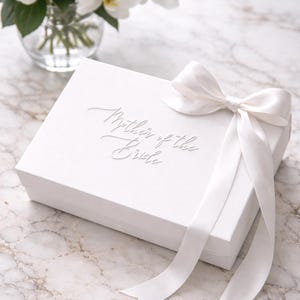 May include: A white gift box, embossed with "Mother of the Bride", is adorned with a satin bow and ribbon. A bouquet of white orchids in a glass vase is in the background. The box rests on a marble surface.