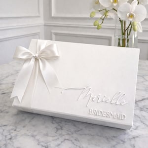 May include: A rectangular white gift box featuring a satin ribbon bow on the left side. The lid is customised with the name Marielle in a raised script font and the word BRIDESMAID in a raised block font, both in a matching white finish.