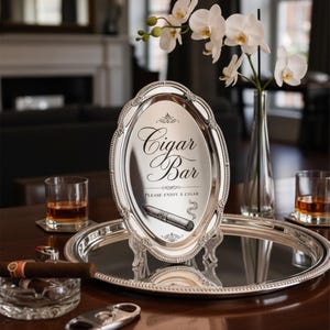 May include: An oval silver-toned tray with a beaded rim displaying the text Cigar Bar and Please Enjoy a Cigar, featuring a graphic of a cigar. The tray sits on a larger round silver-toned tray alongside two glasses of amber liquid and a cigar.