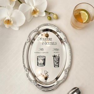May include: An oval silver-toned metal tray with an ornate embossed border. The center features black text reading Signature Cocktails, His Whiskey Ginger, Hers Mojito, and the initials D&R, accompanied by black line-art illustrations of two glasses.