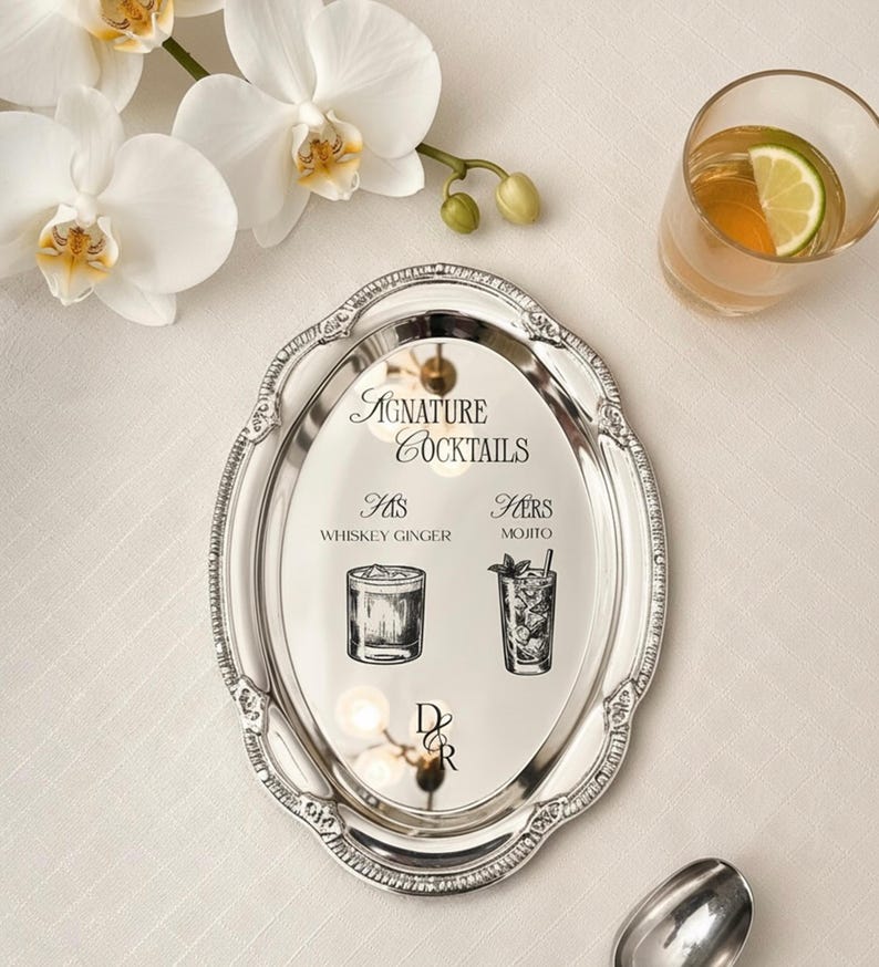 May include: An oval silver-toned metal tray with an ornate embossed border. The center features black text reading Signature Cocktails, His Whiskey Ginger, Hers Mojito, and the initials D&R, accompanied by black line-art illustrations of two glasses.