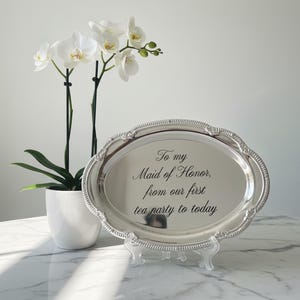 May include: An oval silver-toned serving tray with a decorative beaded rim, displayed on a clear stand. The center of the tray features the engraved black text: To my Maid of Honor, from our first tea party to today. A white orchid plant is nearby.