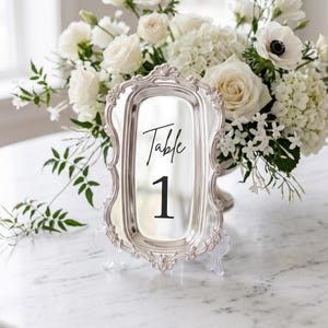 May include: A rectangular silver-toned table number sign with an ornate, decorative border and a reflective center surface. The sign features the black printed text "Table 1" and is supported by a clear acrylic stand, displaying a polished finish.