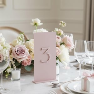 May include: A pale pink table number 3 stands on a marble table, surrounded by floral arrangements of white and pink roses. The table is set with silverware, plates, and wine glasses, suggesting a wedding or event setting.