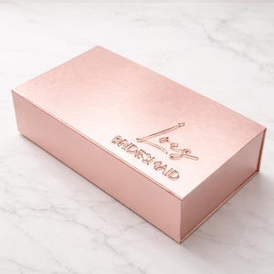 Pu&ograve; includere: Una scatola rettangolare color oro rosa con scritta a specchio color oro rosa "Bridesmaid". La scatola ha una superficie strutturata. La scatola &egrave; su una superficie di marmo bianco.