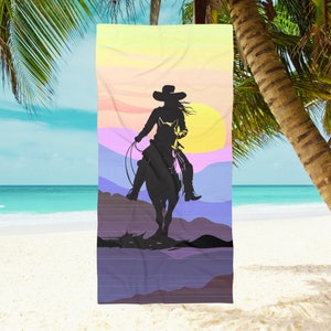 Op de afbeelding: Een strandhanddoek met een silhouet van een cowgirl die op een paard rijdt tegen een kleurrijke zonsondergang.