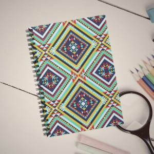 May include: A spiral bound notebook with a colorful geometric pattern featuring blue, red, yellow, and white squares and triangles. The notebook is lying on a white surface.