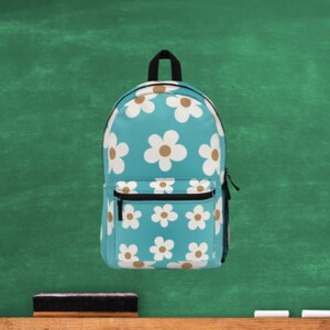 May include: A teal backpack featuring a pattern of white and tan daisies. It has a black zip, a small front pocket, and a black handle. The background is a green chalkboard.