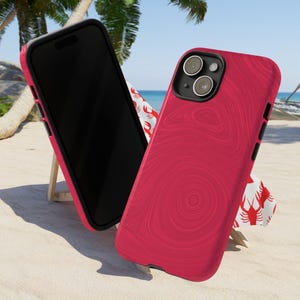 May include: Two red phone cases with a wood grain pattern. One case is on a wooden stand and the other is lying on a white beach towel with red crabs printed on it.