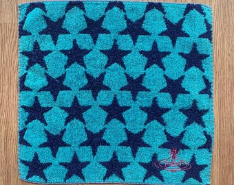 May include: Square turquoise towel with a repeating pattern of navy blue stars. The lower right corner features a pink logo. The towel is made of a soft, absorbent material, perfect for use in a bathroom or kitchen.