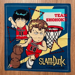 May include: A blue square handkerchief featuring a basketball theme. The design includes two cartoon figures in red basketball uniforms, a basketball hoop, and the text "TEAM SHOHOKU" and "SLAM DUNK".