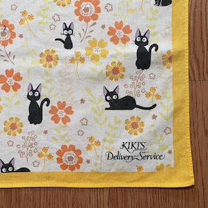 May include: A white square cloth with a yellow border featuring a repeating pattern of black cats, orange and yellow flowers, and green leaves. The text "KIKIS Delivery Service" is printed in black.
