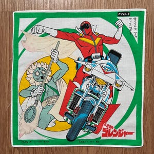 Puede incluir: Un pañuelo de algodón vintage con un personaje de superhéroe rojo y blanco montando una motocicleta. El superhéroe lleva un traje rojo con una capa amarilla y un casco blanco. La motocicleta es blanca con una franja roja. El pañuelo tiene un borde verde y un fondo amarillo con un personaje verde y blanco en el fondo. El texto "パイロット せくらんぼくみ はっそつゃ コレンジャー" está impreso en el pañuelo.