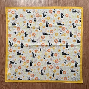 May include: A square, white handkerchief with a repeating pattern of black cat illustrations and orange and yellow flowers. The handkerchief has a yellow border and the text "KIKI'S Delivery Service" in the bottom right corner.