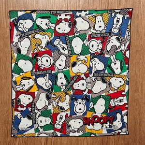 May include: A square Snoopy patterned bandana with a patchwork design. The design features Snoopy in various poses, set against red, yellow, green, and blue backgrounds. The words "THE SUPERBEAGLE" and "SNOOPY" are also visible.