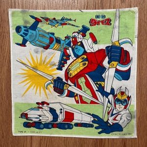 May include: A vintage, square, light green and white cloth featuring colorful illustrations of Japanese anime characters. The design includes a robot, a spaceship, and a character with a helmet, all in vibrant colors. The cloth has Japanese text.