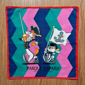 May include: A red, blue and pink bandana with a zig-zag pattern. The bandana features two cartoon characters, a dog and a rabbit, holding flags with anchors. The text "Parque España" is printed on the bandana.