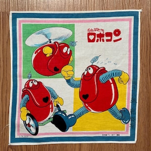 Puede incluir: Un pañuelo de algodón blanco con un borde azul, verde, rosa y amarillo. En el centro, tres robots rojos con ojos negros y un corazón en el pecho. El texto "ガンバレ!! ロボコン" está impreso en negro en la esquina superior derecha del pañuelo.