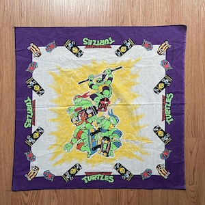 Könnte beinhalten: Lilafarbener Bandana mit einem weißen Zentrum, auf dem die Teenage Mutant Ninja Turtles beim Skateboardfahren zu sehen sind. Der Bandana hat die Worte "Teenage Mutant Ninja Turtles" und "Turtles" aufgedruckt.