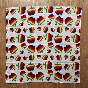 May include: A white cotton bandana with a repeating pattern of colorful rainbow apples. The bandana has a small tag with the text "K.O.A. 100%".