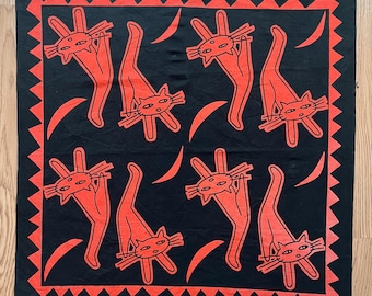 Może przedstawiać: Czarna kwadratowa bandana z powtarzającym się wzorem stylizowanych pomarańczowych kotów i półksiężyców. Bandana ma czerwony geometryczny obramowanie. Koty są przedstawione z długimi ciałami i wyciągniętymi łapami.