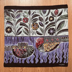 Puede incluir: Textil cuadrado con borde negro con un diseño estilizado. La mitad superior tiene patrones florales y de hojas en blanco, negro y varios colores. La mitad inferior representa dos peces en tonos rojos, amarillos y negros, sobre líneas onduladas azules.