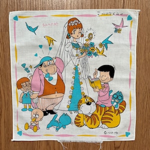 Puede incluir: Un pañuelo blanco con una ilustración colorida de una escena de boda. La novia lleva un vestido azul y blanco y un velo. El novio lleva un traje rosa y un sombrero azul. Hay varios otros personajes en la escena, incluyendo un gato, un perro y un pájaro. El texto "フクオカ" está impreso en la esquina inferior derecha.