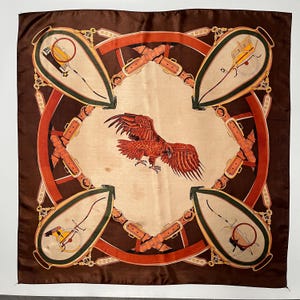 Puede incluir: Un pañuelo cuadrado de seda con una ilustración central de un águila marrón en vuelo. El diseño incluye una disposición circular de cinturones y hebillas de cuero naranja, con cuatro motivos en forma de escudo en las esquinas con carruajes.